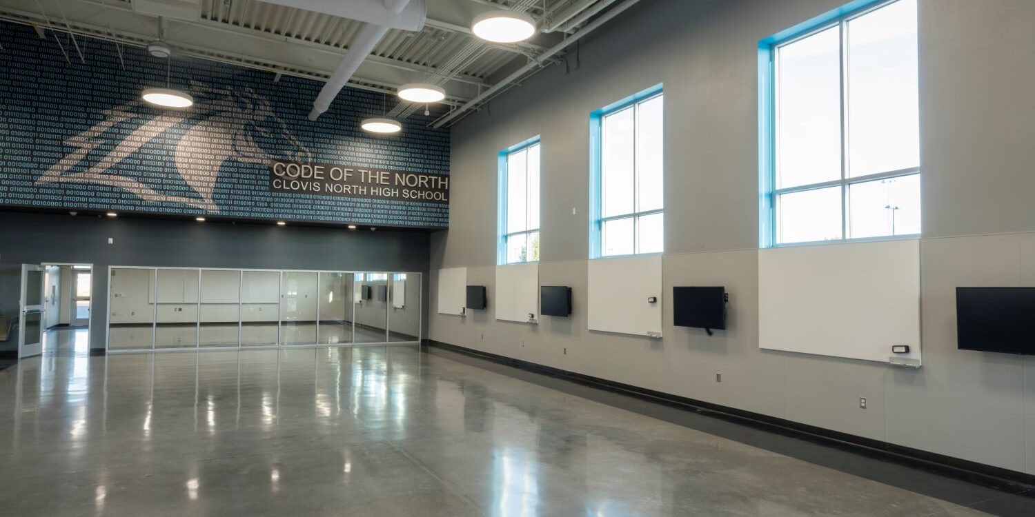 A professional, indoor photo of a large, empty, modern common room or multi-purpose room at Clovis North High School. The walls are a light gray with large windows framed in white. A large mural featuring a horse made of binary code with the words "CODE OF THE NORTH, CLOVIS NORTH HIGH SCHOOL" is painted on one wall. The floor is a polished concrete, and the ceiling has exposed ducts and modern light fixtures. A wall of mirrors is visible in the background, to the left.