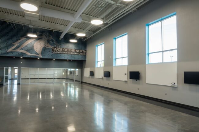 A professional, indoor photo of a large, empty, modern common room or multi-purpose room at Clovis North High School. The walls are a light gray with large windows framed in white. A large mural featuring a horse made of binary code with the words "CODE OF THE NORTH, CLOVIS NORTH HIGH SCHOOL" is painted on one wall. The floor is a polished concrete, and the ceiling has exposed ducts and modern light fixtures. A wall of mirrors is visible in the background, to the left.