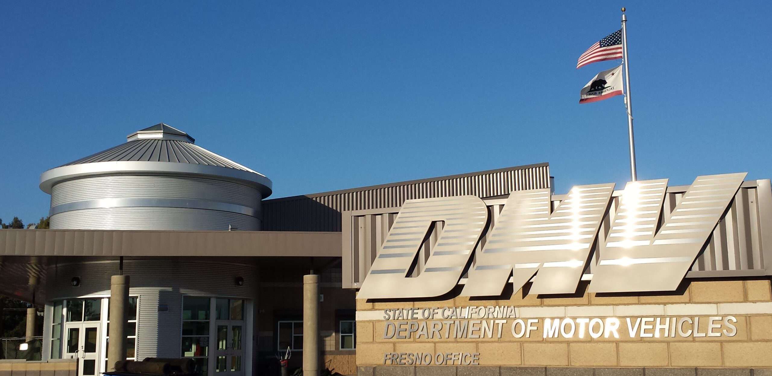 A close-up, outdoor shot of the front of a modern DMV building. The building has a large, metallic, stylized "DMV" sign. Below it, the text "STATE OF CALIFORNIA DEPARTMENT OF MOTOR VEHICLES FRESNO OFFICE" is visible. A circular, metallic section of the building with a dome roof is visible to the left, and a flagpole with the American and California state flags is visible to the right. The sky is a clear blue.