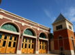 A low-angle, outdoor shot of the front of a modern fire station in Clovis. The building has a red brick facade with large, arched, wooden garage doors and "CLOVIS FIRE STATION" spelled out in green letters above the doors. To the right, a tall, orange and yellow tower with a pointed roof rises into a clear blue sky with some wispy clouds.