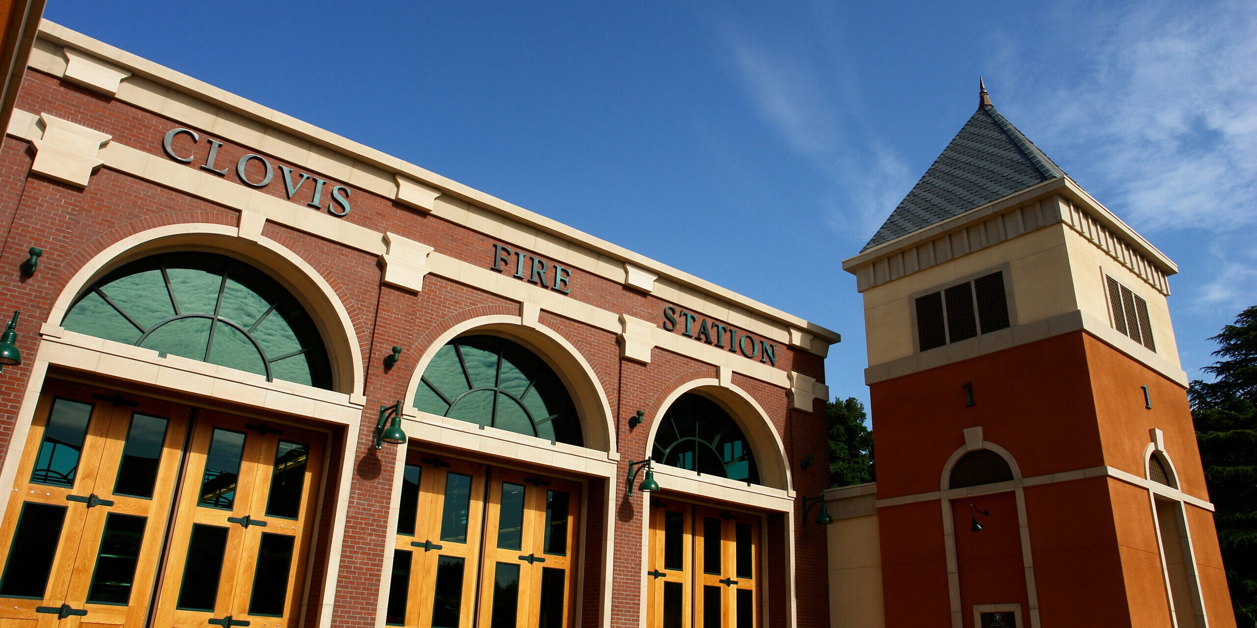CLOVIS FIRE STATION A low-angle, outdoor shot of the front of a modern fire station in Clovis. The building has a red brick facade with large, arched, wooden garage doors and "CLOVIS FIRE STATION" spelled out in green letters above the doors. To the right, a tall, orange and yellow tower with a pointed roof rises into a clear blue sky with some wispy clouds.