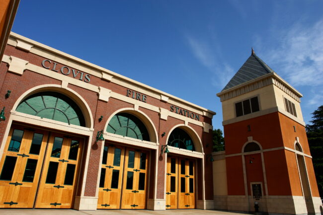 A low-angle, outdoor shot of the front of a modern fire station in Clovis. The building has a red brick facade with large, arched, wooden garage doors and "CLOVIS FIRE STATION" spelled out in green letters above the doors. To the right, a tall, orange and yellow tower with a pointed roof rises into a clear blue sky with some wispy clouds.