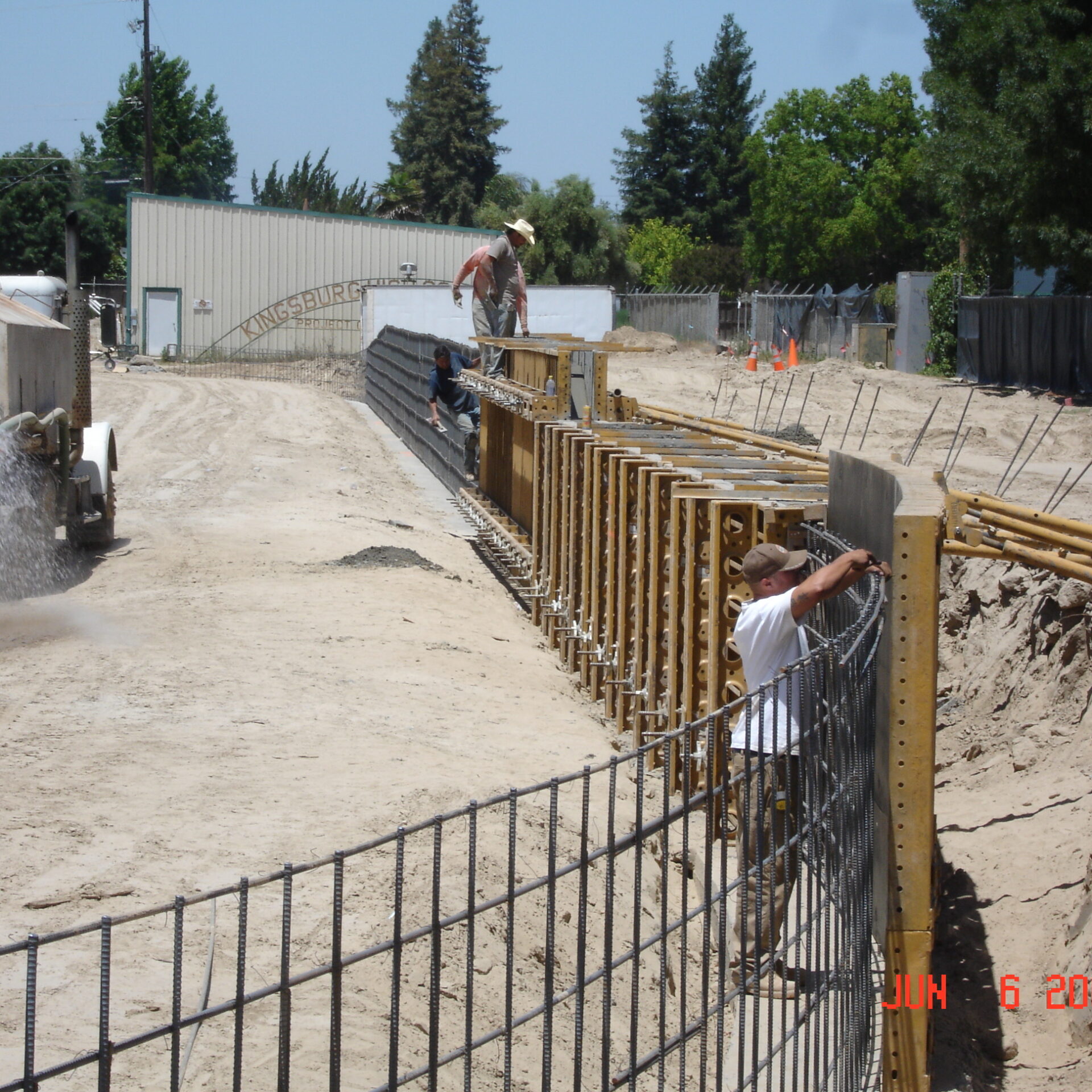 A ground-level, daytime shot of a construction site. The image shows a long trench with wooden forms and rebar cages in place, indicating preparation for a concrete foundation or retaining wall. Several construction workers are visible, and in the background, a white cement truck is spraying water onto the ground, creating a mist. The ground is a mix of dirt and sand. In the distance, trees and a large metal building are visible. The image has a date stamp of "JUN 6 2005" in the bottom right corner.