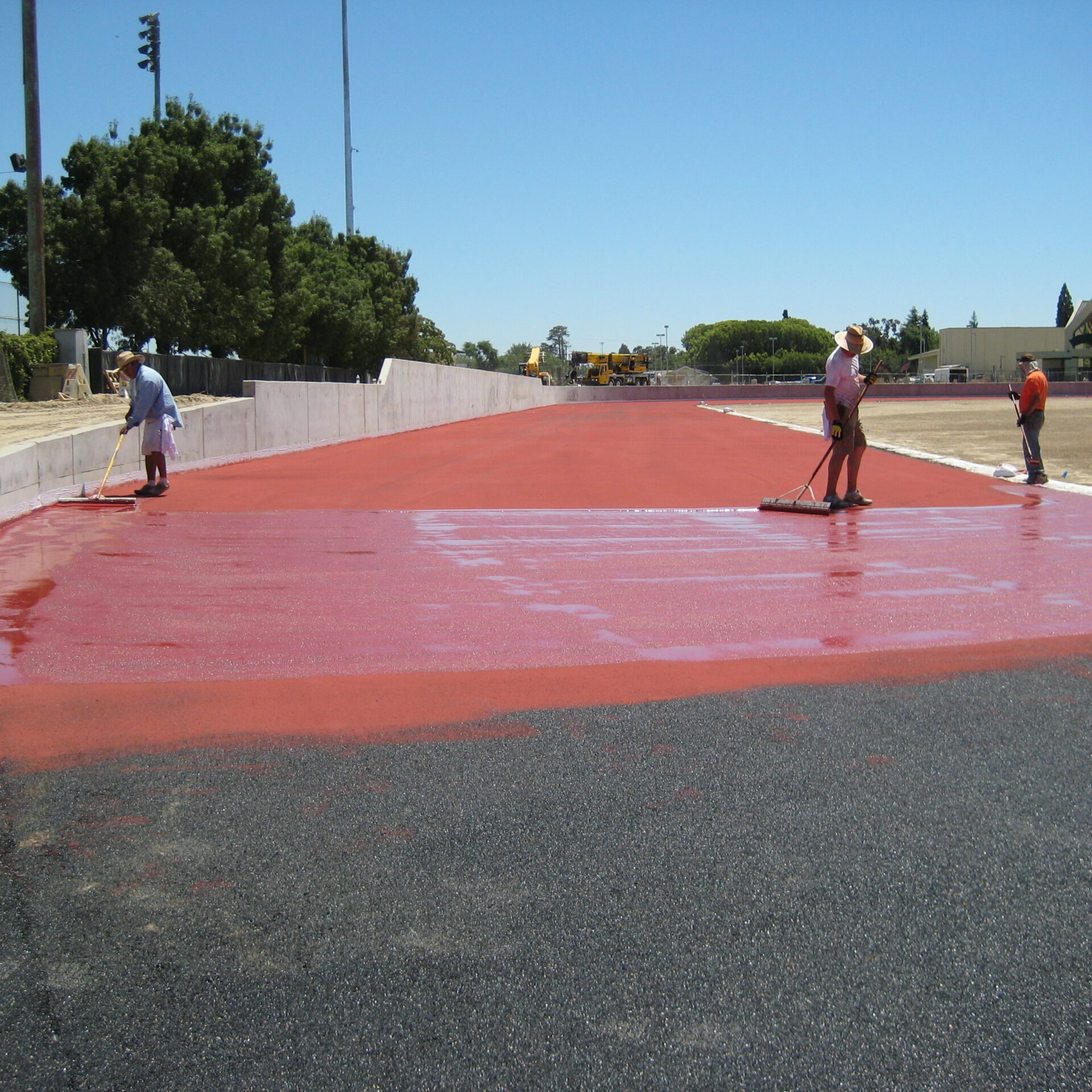 A daytime, ground-level shot of three construction workers applying a red sealant or coating to a running track surface at a high school stadium. The workers are using long-handled squeegees and rollers to smooth the liquid onto the black asphalt base. The track is adjacent to a white concrete wall. In the background, there is a sports field and a large green tree. The sky is a clear, bright blue. The scene captures a phase of the sports facility's construction.