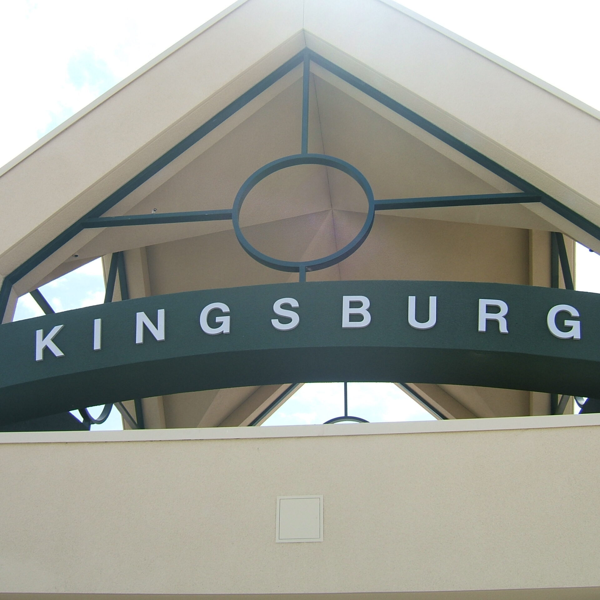A low-angle, close-up shot of the entrance facade of the Kingsburg High School football stadium. The main feature is the word "KINGSBURG" spelled out in white block letters on a dark green banner that curves downward. The building has a peaked roof with a light tan stucco finish and a large circular window opening. The image focuses on the architectural details of the entrance, with a cloudy blue sky visible through the opening and in the background.