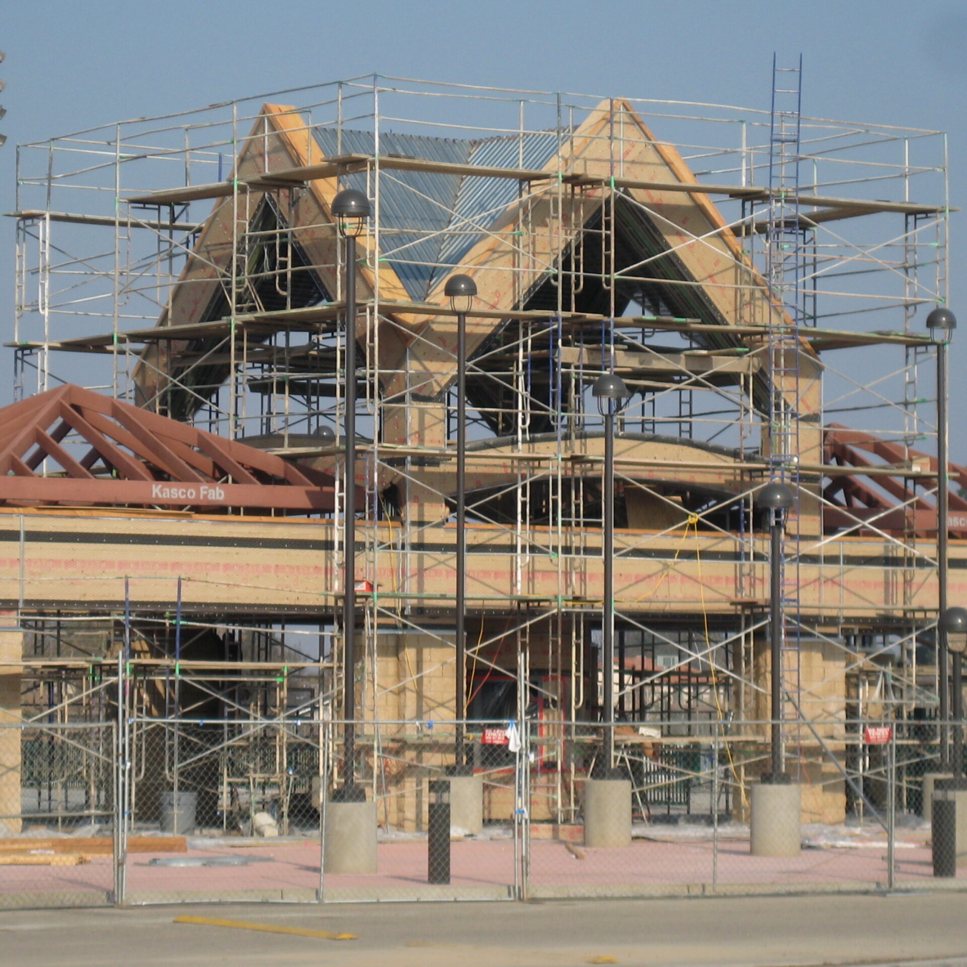 A wide-angle, exterior shot of the Kingsburg High School football stadium entrance under construction. The structure is surrounded by a dense network of metal scaffolding, with a chain-link fence in the foreground. The building has a large, unfinished peaked roof structure and a tan stone facade. The roof's wooden frame is visible, and some red roofing material is on the smaller side roofs. Street lamps with multiple lights are placed in the foreground. The sky is a clear blue. The overall image gives a sense of a large construction project in progress.