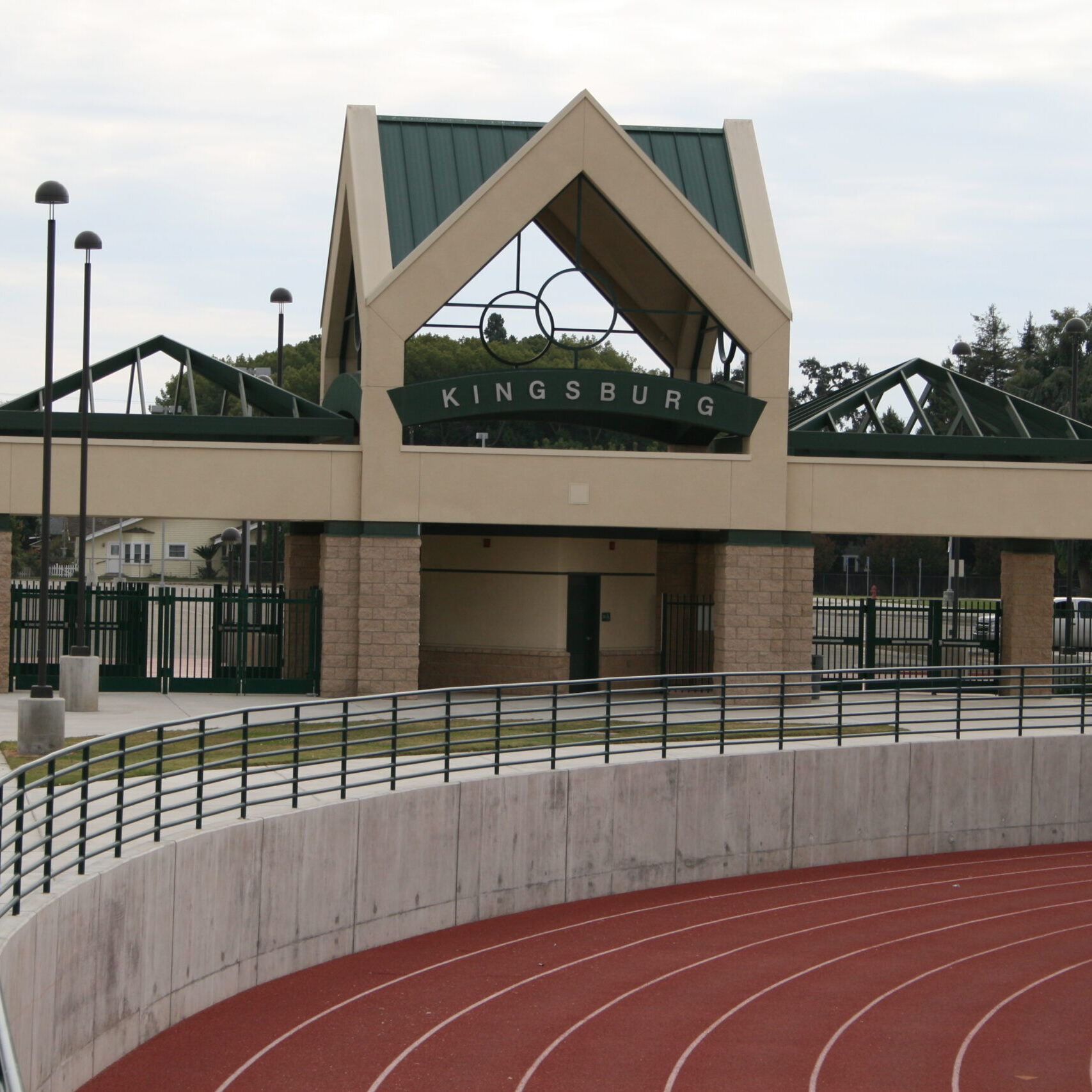 A medium-angle exterior shot of the entrance to a sports stadium at Kingsburg High School. The entrance is a two-story structure with a peaked green roof and a tan stone facade. The word "KINGSBURG" is visible in large letters above the archway. A concrete track with red lanes is in the foreground, curving to the right, with a black metal railing separating it from the paved concourse area. The sky is overcast. In the background, there are residential buildings and trees. The overall image captures the entrance to a well-maintained athletic facility.