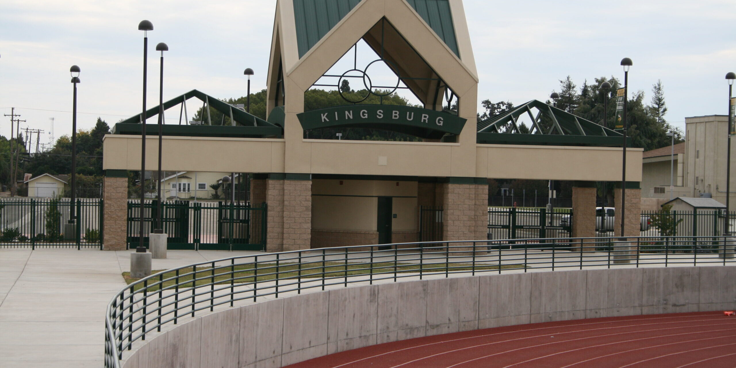 A medium-angle exterior shot of the entrance to a sports stadium at Kingsburg High School. The entrance is a two-story structure with a peaked green roof and a tan stone facade. The word "KINGSBURG" is visible in large letters above the archway. A concrete track with red lanes is in the foreground, curving to the right, with a black metal railing separating it from the paved concourse area. The sky is overcast. In the background, there are residential buildings and trees. The overall image captures the entrance to a well-maintained athletic facility.
