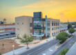 A professional, outdoor photo of a multi-story, modern building with tan and red facades and a prominent glass-enclosed section. The building is located next to a street with a few trees and streetlights, and a red brick building with a tower is visible in the background. The photo is taken at sunset, with a vibrant orange and yellow sky.