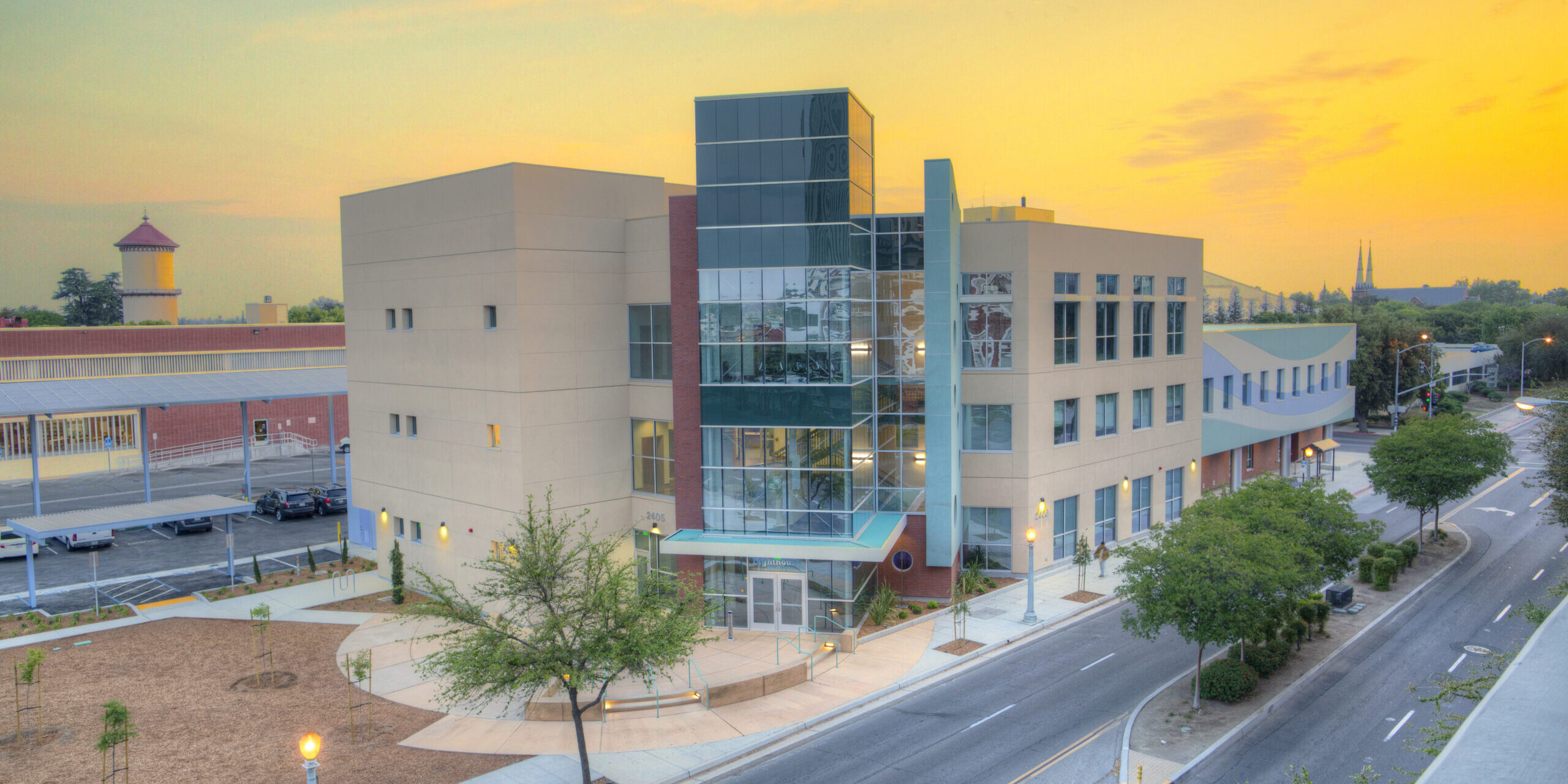 A professional, outdoor photo of a multi-story, modern building with tan and red facades and a prominent glass-enclosed section. The building is located next to a street with a few trees and streetlights, and a red brick building with a tower is visible in the background. The photo is taken at sunset, with a vibrant orange and yellow sky.