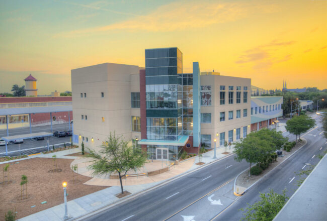 A professional, outdoor photo of a multi-story, modern building with tan and red facades and a prominent glass-enclosed section. The building is located next to a street with a few trees and streetlights, and a red brick building with a tower is visible in the background. The photo is taken at sunset, with a vibrant orange and yellow sky.