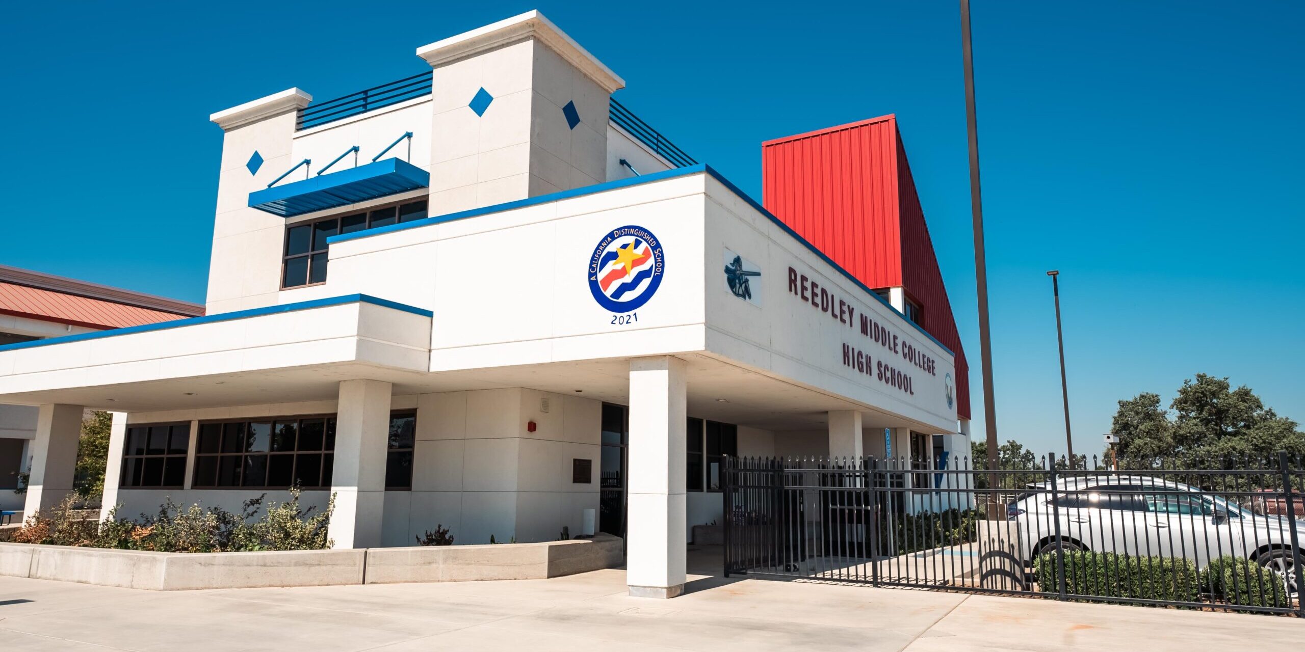 A professional, wide-angle, outdoor photo of the front of a modern high school building. The building has a white stucco exterior with blue trim and a red metal roof section. The school name, "REEDLEY MIDDLE COLLEGE HIGH SCHOOL," is visible on the facade. A circular logo with a wave design and the year "2021" is also visible. The entrance is covered by a blue awning, and a black fence runs along the front of the building. The sky is a clear, bright blue.