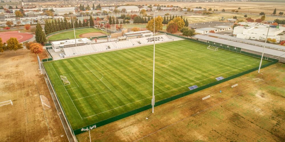 The Clovis East Soccer Stadium An aerial view of a large, well-maintained sports field, which is the Clovis East Soccer Stadium. The field is covered in bright green grass, with white lines marking a soccer pitch. A set of white bleachers sits on one side of the field, and a baseball field is visible behind it. The stadium is surrounded by a brown field, and in the background, a cluster of residential and commercial buildings are visible under a hazy sky. A row of solar panels is visible on the roof of a building adjacent to the field.
