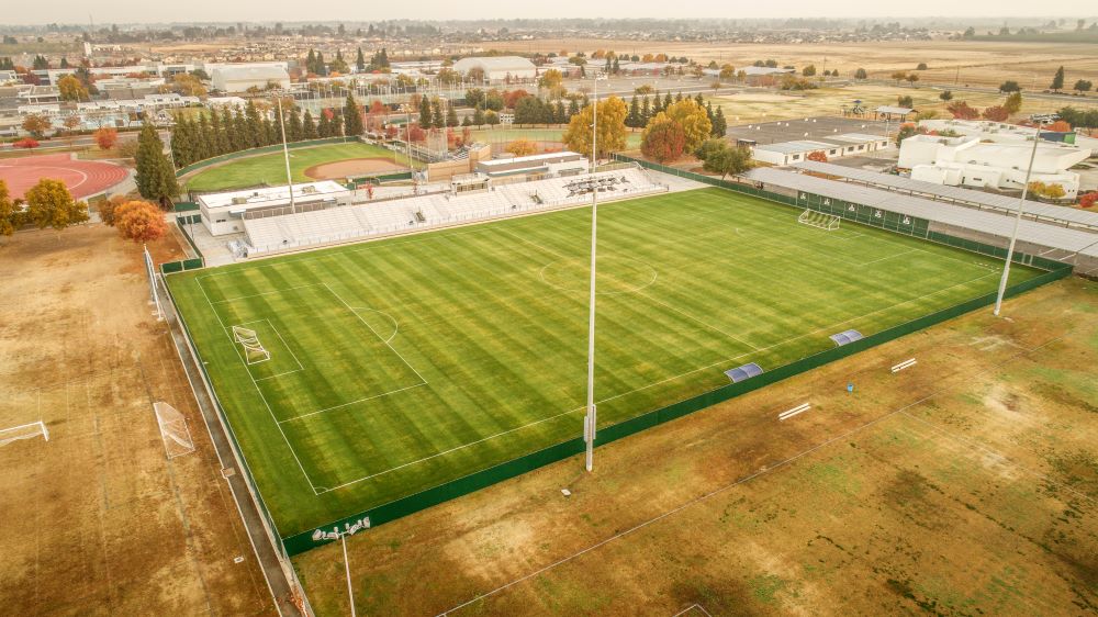 An aerial view of a large, well-maintained sports field, which is the Clovis East Soccer Stadium. The field is covered in bright green grass, with white lines marking a soccer pitch. A set of white bleachers sits on one side of the field, and a baseball field is visible behind it. The stadium is surrounded by a brown field, and in the background, a cluster of residential and commercial buildings are visible under a hazy sky. A row of solar panels is visible on the roof of a building adjacent to the field.