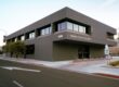A wide-angle, outdoor shot of a modern, two-story commercial building, which is the Fresno County Recorder's office. The building is a dark gray color with large, dark-tinted windows. The address, "1250", and the name "FRESNO COUNTY RECORDER" are visible on the facade. The photo is taken from a street corner, showing the building's main entrance and a sidewalk with a street sign that says "ENTER ONLY." The sky is a light blue, and a tree is visible on the left.