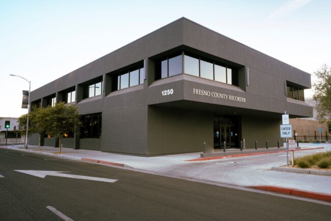 A wide-angle, outdoor shot of a modern, two-story commercial building, which is the Fresno County Recorder's office. The building is a dark gray color with large, dark-tinted windows. The address, "1250", and the name "FRESNO COUNTY RECORDER" are visible on the facade. The photo is taken from a street corner, showing the building's main entrance and a sidewalk with a street sign that says "ENTER ONLY." The sky is a light blue, and a tree is visible on the left.