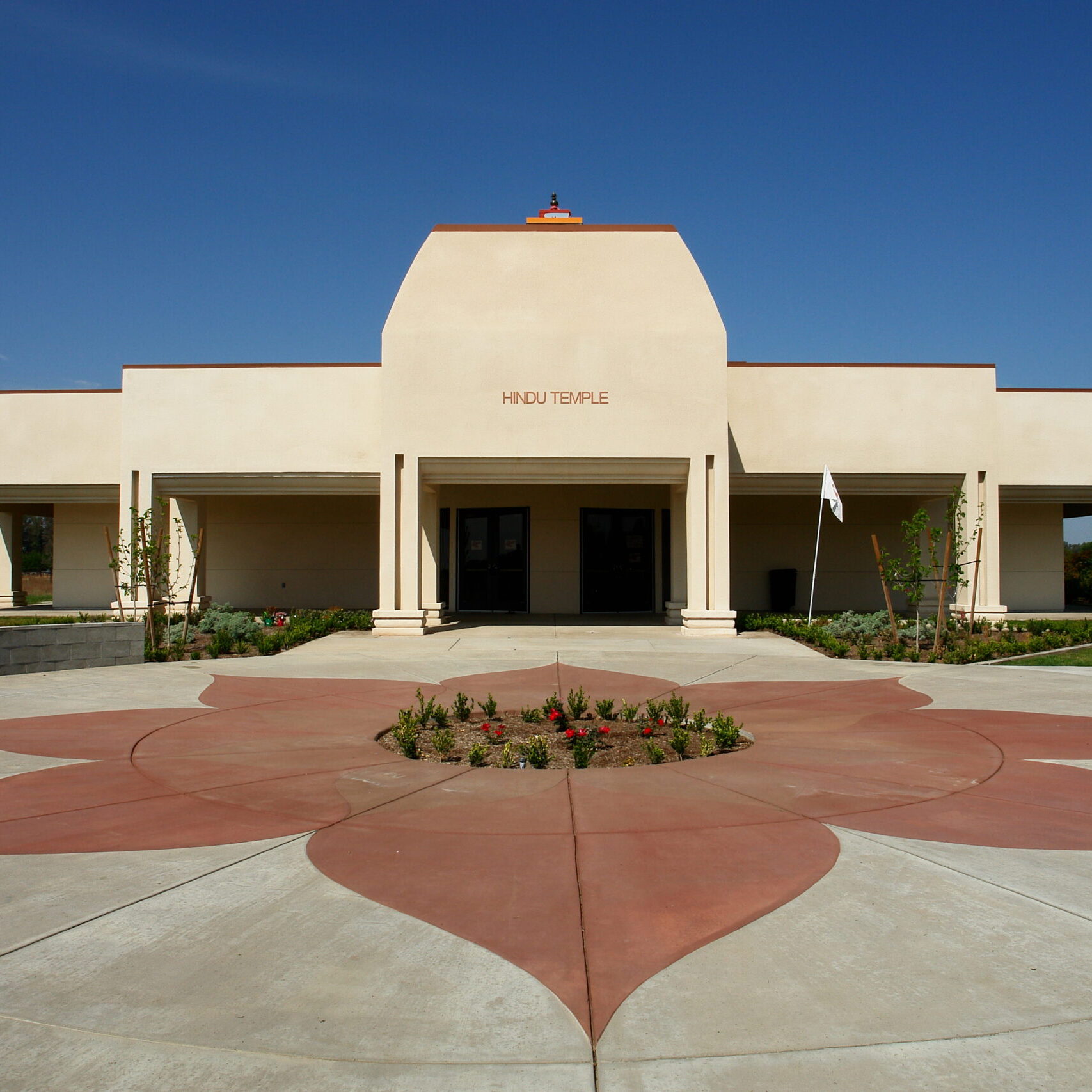 A symmetrical, ground-level exterior shot of a modern Hindu temple building. The facade is a light-colored stucco with a large, elevated central section that has the words "HINDU TEMPLE" on it. A covered portico with a series of pillars extends from the main building. The foreground features a decorative red concrete star or flower pattern embedded in the gray concrete walkway. A small, landscaped garden is in the center of the star pattern. The sky is a clear blue.