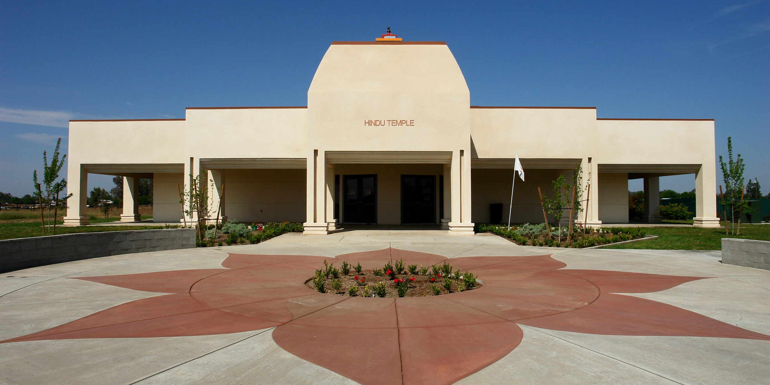 A symmetrical, ground-level exterior shot of a modern Hindu temple building. The facade is a light-colored stucco with a large, elevated central section that has the words "HINDU TEMPLE" on it. A covered portico with a series of pillars extends from the main building. The foreground features a decorative red concrete star or flower pattern embedded in the gray concrete walkway. A small, landscaped garden is in the center of the star pattern. The sky is a clear blue.