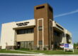 A slightly low-angle, exterior shot of a modern, two-story commercial building on a sunny day. The building's facade is a mix of light-colored stucco, a darker tan central tower, and a stone veneer base. The words "Ear, Allergy, & Sinus Center" are visible on the left side of the building. In the foreground, there is a manicured lawn and two "For Lease/Sale" signs for Colliers Tingey. The sky is a clear, deep blue. The image captures the building from a street-level perspective, highlighting its clean lines and professional appearance.