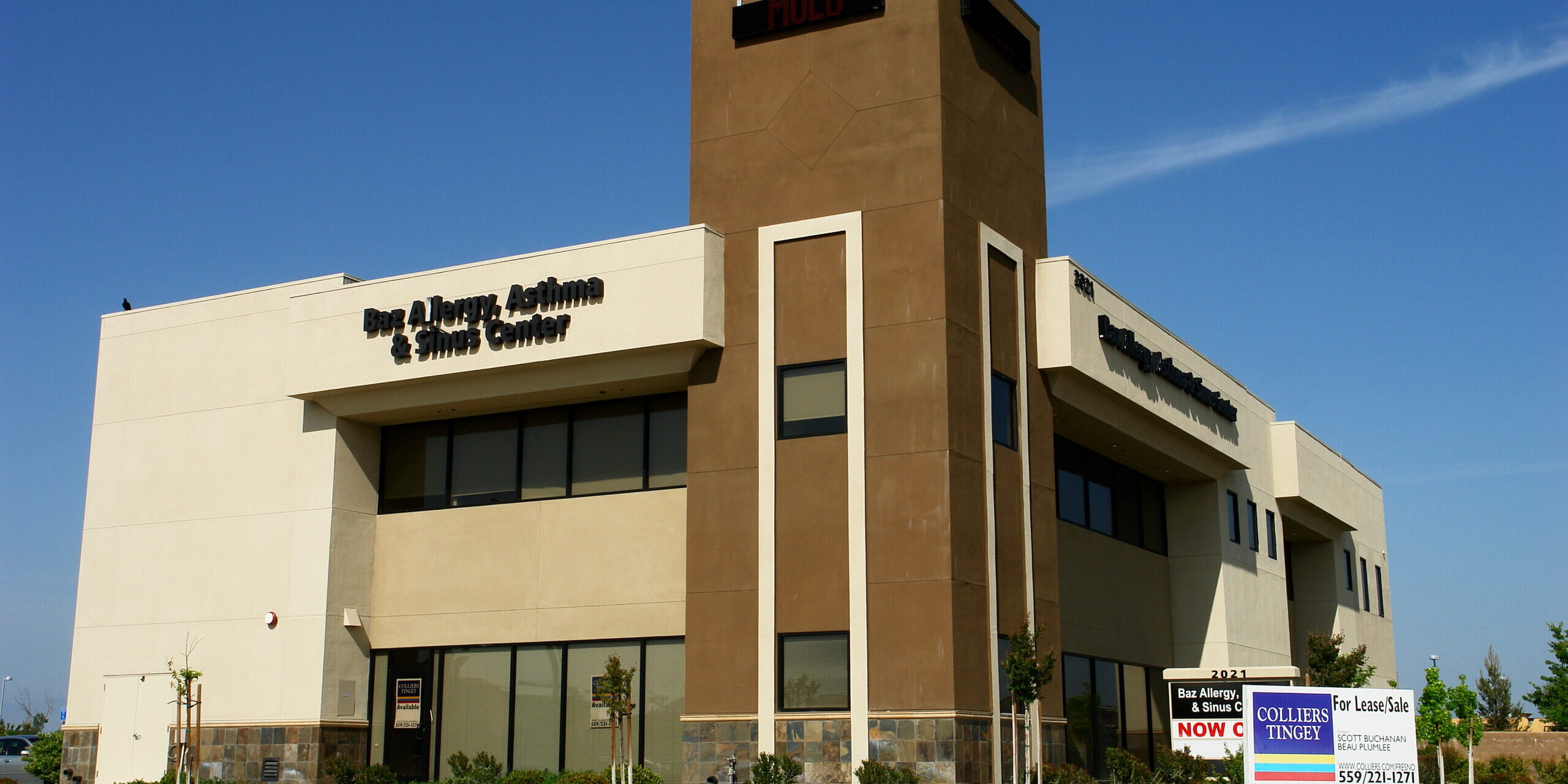 A slightly low-angle, exterior shot of a modern, two-story commercial building on a sunny day. The building's facade is a mix of light-colored stucco, a darker tan central tower, and a stone veneer base. The words "Ear, Allergy, & Sinus Center" are visible on the left side of the building. In the foreground, there is a manicured lawn and two "For Lease/Sale" signs for Colliers Tingey. The sky is a clear, deep blue. The image captures the building from a street-level perspective, highlighting its clean lines and professional appearance.