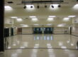 A symmetrical, interior shot of a large, empty multi-purpose room at Oakhurst Elementary School. The room has a highly polished, light-colored floor that reflects the ceiling lights and the double doors at the far end. The walls are a light tan color with a wood trim. Above, the ceiling is a mix of acoustic tiles with square fluorescent lights and a large set of theater spotlights. There are two sets of double doors with "EXIT" signs above them on the back wall. The room appears to be ready for an event or activity.