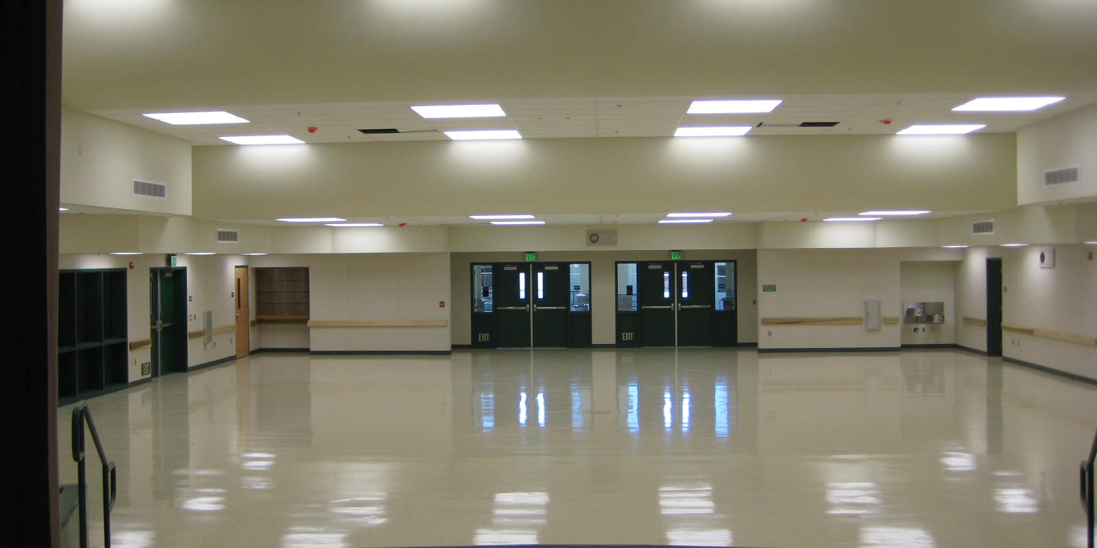 Oakhurst Elementary New Multi Purpose Building (13) A symmetrical, interior shot of a large, empty multi-purpose room at Oakhurst Elementary School. The room has a highly polished, light-colored floor that reflects the ceiling lights and the double doors at the far end. The walls are a light tan color with a wood trim. Above, the ceiling is a mix of acoustic tiles with square fluorescent lights and a large set of theater spotlights. There are two sets of double doors with "EXIT" signs above them on the back wall. The room appears to be ready for an event or activity.