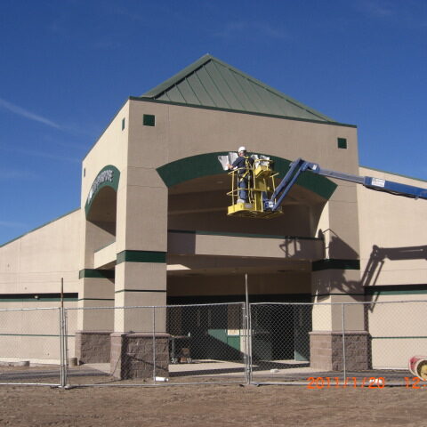 A wide-angle, exterior shot of a building under construction, likely a school or commercial building. The building has a light-colored stucco facade and a green, sloped metal roof. A blue mechanical lift with a worker in the basket is positioned near the entrance, where the worker is doing work on the facade. The building's entrance has a prominent archway with dark green accents. The foreground is a dirt and gravel area, enclosed by a chain-link fence. The sky is a clear, bright blue. A date stamp in the bottom right corner reads "2011/11/20".