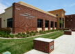 A medium-angle, exterior shot of the Riverdale Community Education Center building on a sunny day. The one-story building has a contemporary design with a prominent red brick section on the front facade and a light-colored stucco section on the right. The words "RIVERDALE COMMUNITY EDUCATION CENTER" and the building number "3160" are visible in relief on the brick wall. A manicured lawn and a landscaped area with plants and flowers line the front of the building. A clean, paved walkway with brick-capped planters is in the foreground. The sky is a bright blue with scattered clouds.