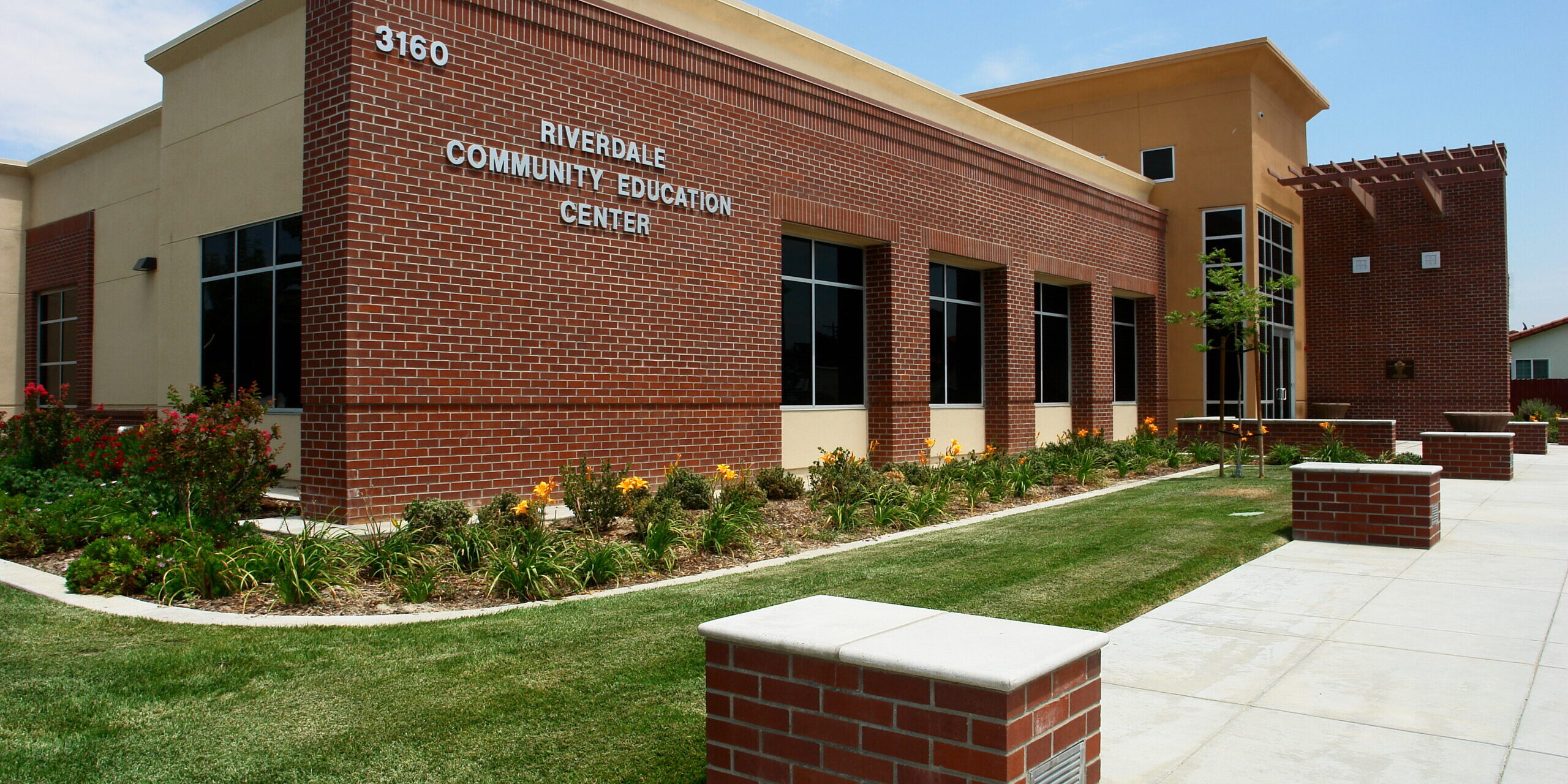 A medium-angle, exterior shot of the Riverdale Community Education Center building on a sunny day. The one-story building has a contemporary design with a prominent red brick section on the front facade and a light-colored stucco section on the right. The words "RIVERDALE COMMUNITY EDUCATION CENTER" and the building number "3160" are visible in relief on the brick wall. A manicured lawn and a landscaped area with plants and flowers line the front of the building. A clean, paved walkway with brick-capped planters is in the foreground. The sky is a bright blue with scattered clouds.