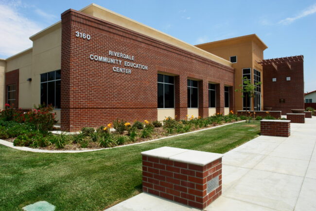 A medium-angle, exterior shot of the Riverdale Community Education Center building on a sunny day. The one-story building has a contemporary design with a prominent red brick section on the front facade and a light-colored stucco section on the right. The words "RIVERDALE COMMUNITY EDUCATION CENTER" and the building number "3160" are visible in relief on the brick wall. A manicured lawn and a landscaped area with plants and flowers line the front of the building. A clean, paved walkway with brick-capped planters is in the foreground. The sky is a bright blue with scattered clouds.
