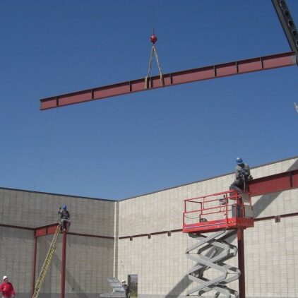 A low-angle, eye-level shot of a building under construction. Two construction workers in hard hats and safety gear are visible. One worker is on a red scissor lift, and another is on a ladder. They are working to guide a long, red steel beam being lowered by a construction crane, which is partially visible at the top right. The building's walls are made of gray concrete blocks. The sky is bright blue and cloudless.