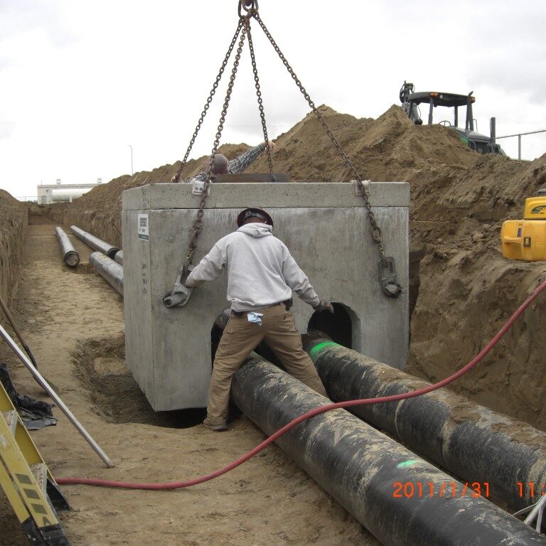 A photograph taken at a construction site shows two workers in a deep trench, guiding a large, pre-cast concrete box as it is lowered by a crane. The box is suspended by heavy-duty chains. One worker, wearing a hard hat, is crouched and guiding the box by its side, while another is on top of the box. Two large, black pipes run horizontally through the trench. In the background, there is a pile of dirt and construction equipment, and the sky is overcast. A timestamp at the bottom right corner of the image reads "2011/1/31 11:46am".