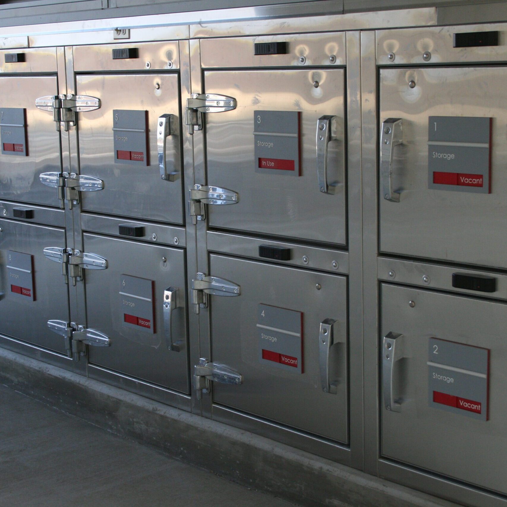 A close-up, eye-level shot of a series of large, stainless steel refrigerated storage compartments. Each unit has a latch handle and a small, gray plaque with a red tag and a number. The plaques read "Storage," "Ride," or "Vacant." The compartments are stacked two high, with an elevated platform in front of them. The entire structure is very clean and reflective.