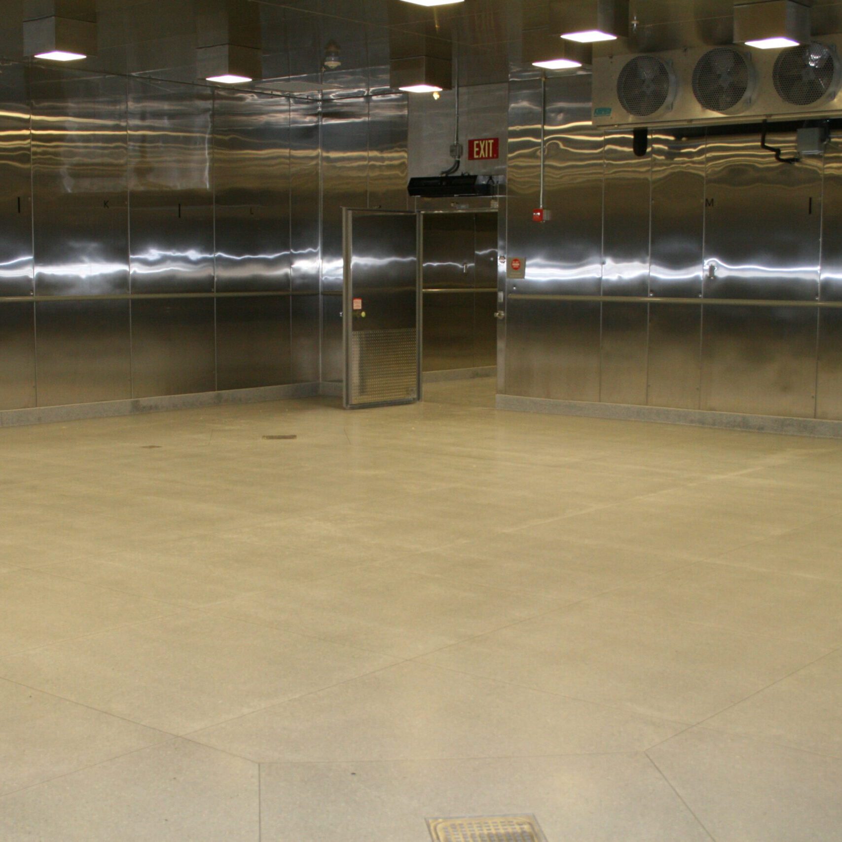 A wide-angle, eye-level shot of a large, empty, stainless steel refrigerated room. The walls are made of reflective, corrugated stainless steel, and the floor is a light-colored tile. In the center of the back wall, there is a large, open steel door with a grated window, showing a dark room beyond. Above the door, a red "Exit" sign is visible. High on the right wall, a bank of four large fans is mounted. The room is lit by several rows of square, recessed lights in the ceiling, creating a ripple effect on the walls.