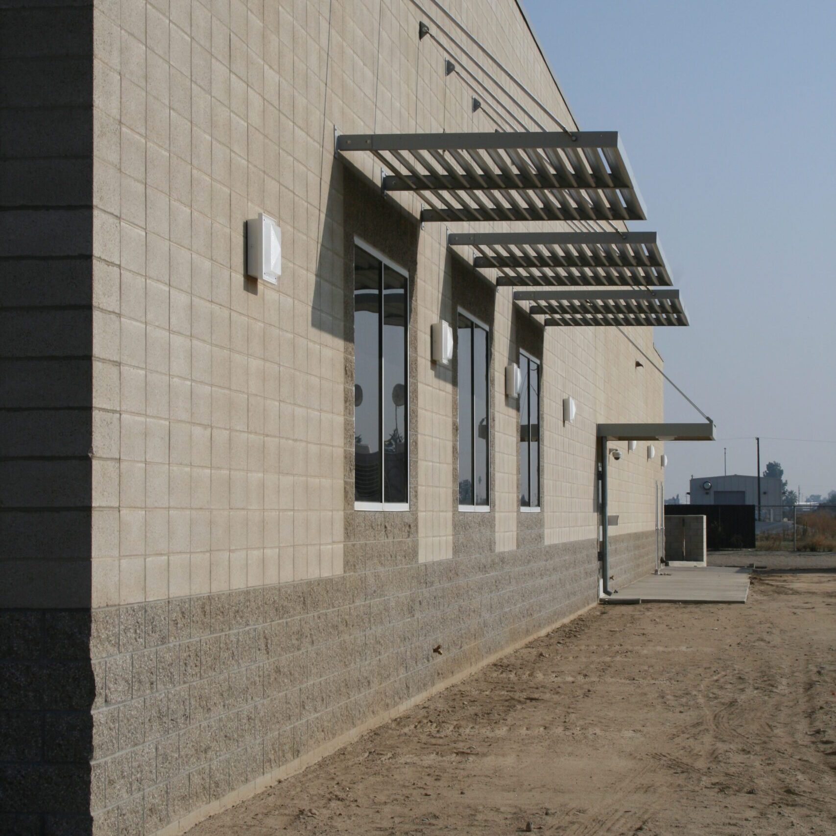 A low-angle, eye-level shot of the side of a large, modern, institutional-style building. The walls are made of two different-colored concrete blocks, a lighter beige color on top and a darker gray on the bottom. Several rectangular windows with gray metal canopies are visible. The sun casts long shadows, and the foreground is a dirt and gravel lot.