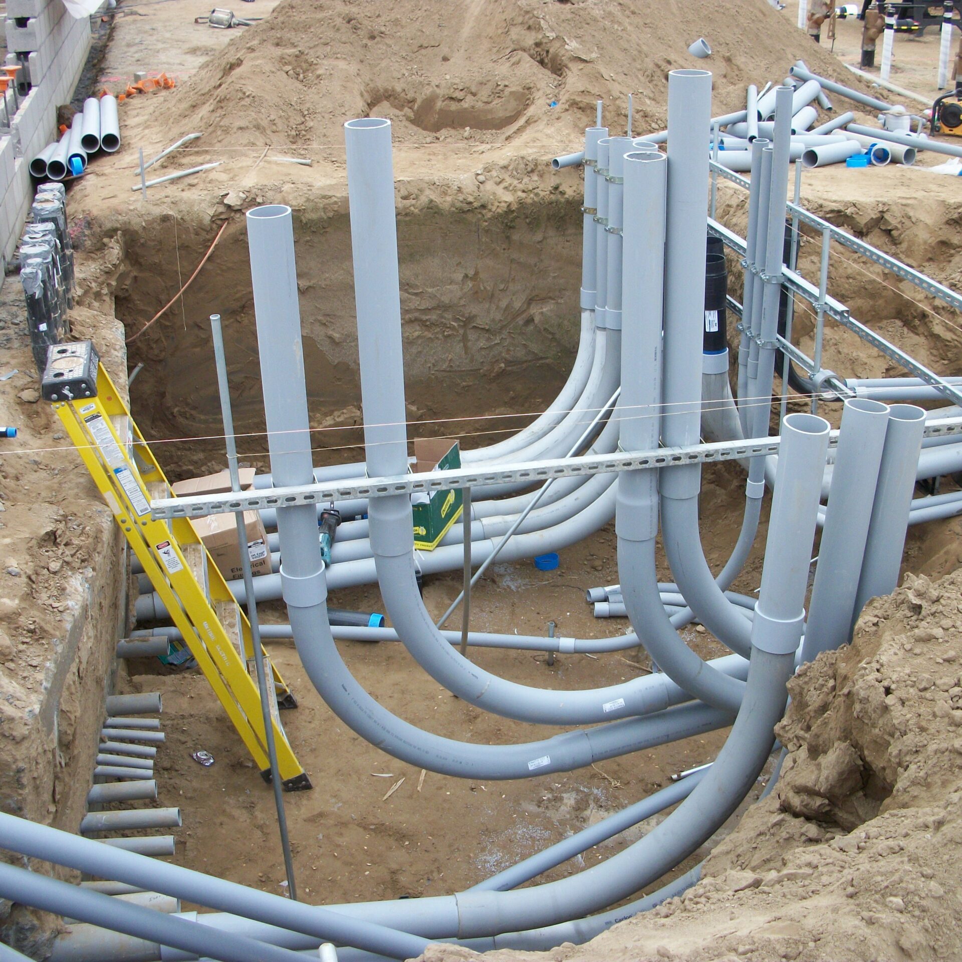 A close-up, eye-level shot of a deep trench on a construction site, revealing an intricate network of gray and blue PVC pipes. The pipes emerge from the ground, bend in various directions, and connect to form large bundles. The surrounding dirt is sandy and light-brown. A yellow ladder is placed in the trench, and various construction tools and materials, including more pipes, are scattered nearby.