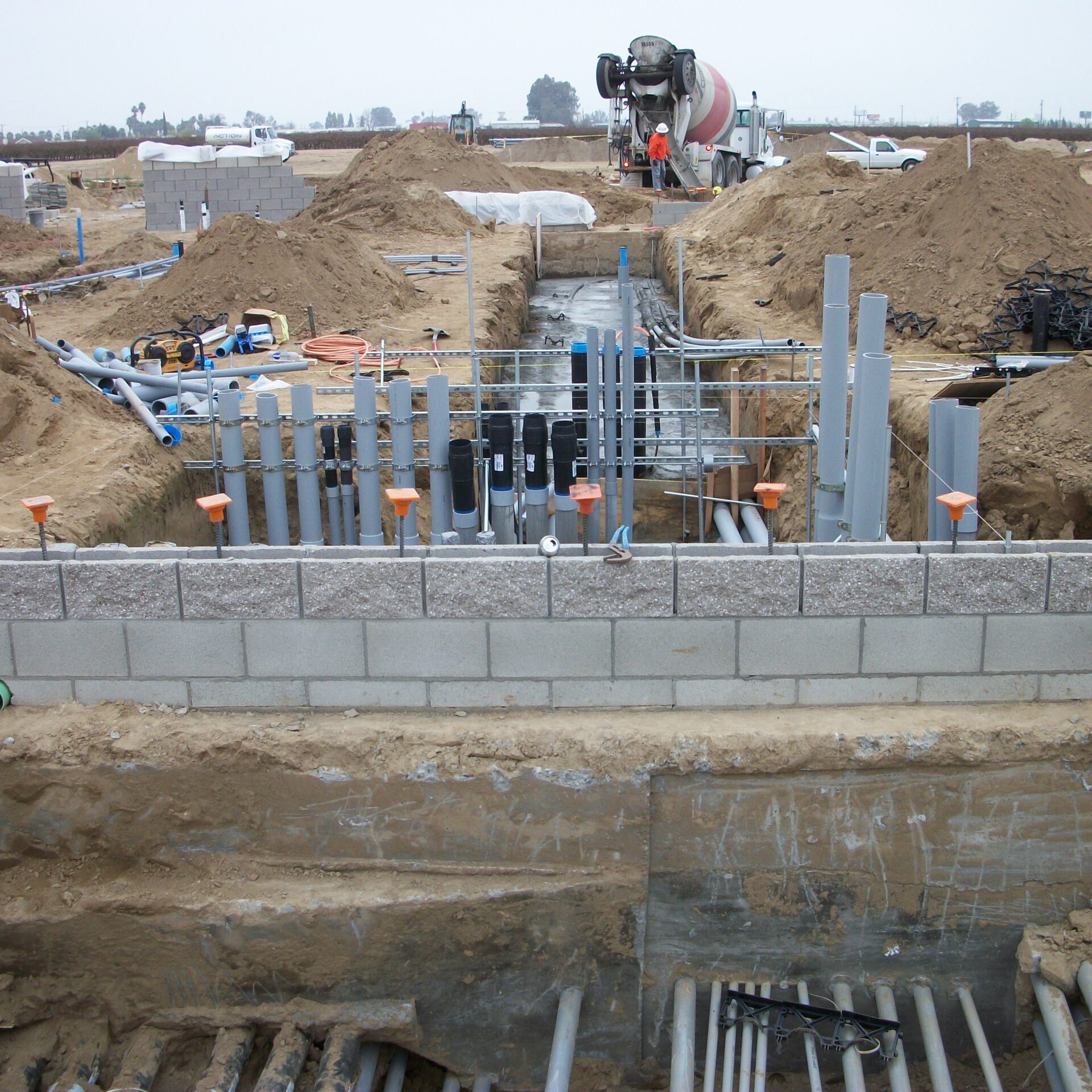 A wide-angle, slightly elevated shot of a construction site showing a deep trench with extensive plumbing and electrical conduit. In the foreground, there is a gray cinder block wall. The trench is surrounded by piles of dirt, and a cement mixer truck is visible in the background, along with a worker in an orange vest. The sky is gray and overcast.