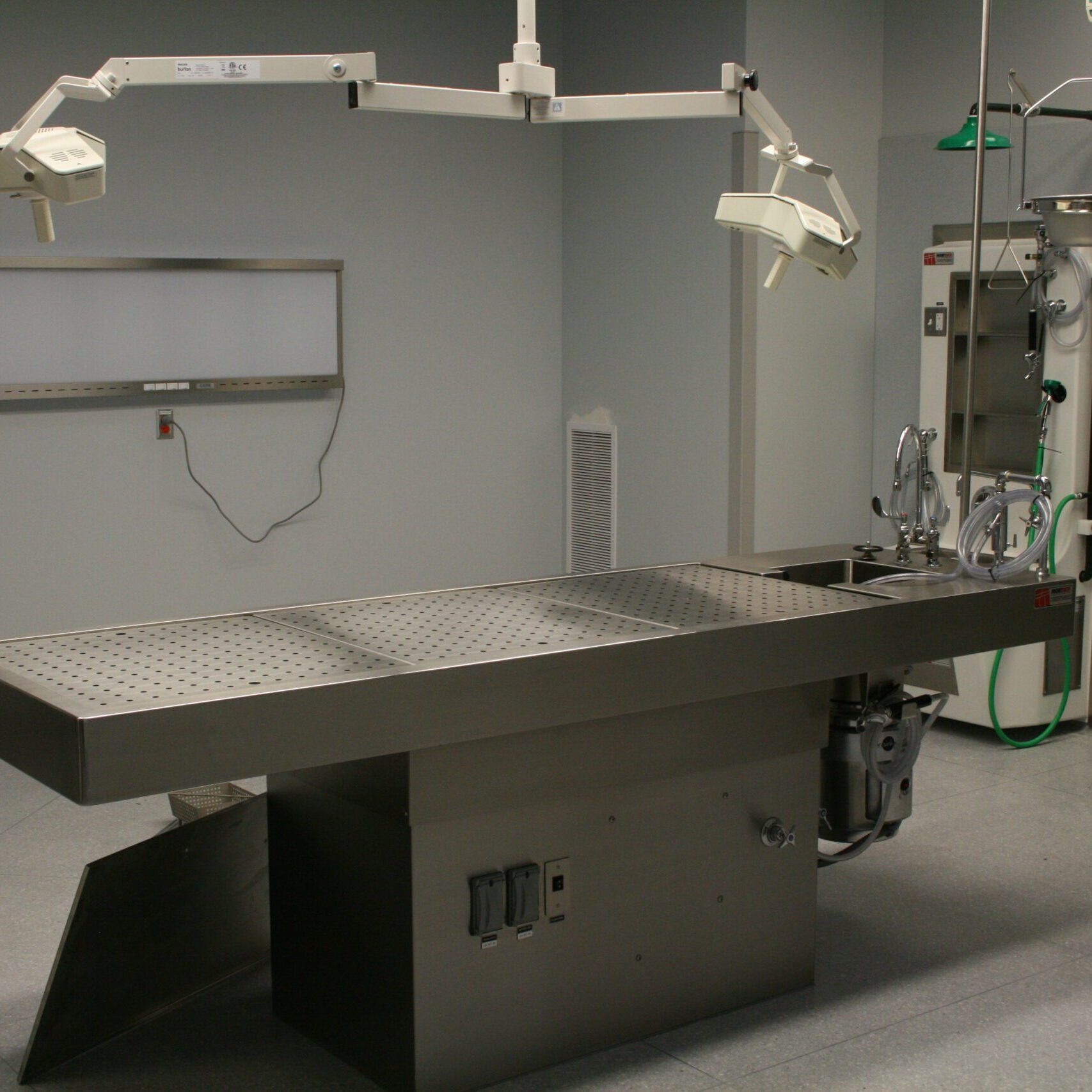 A low-angle, eye-level shot of a clean, modern medical or laboratory room. A large, stainless steel autopsy table with a perforated surface is in the center of the room. Two adjustable surgical lights are suspended from the ceiling above the table. A control panel is built into the base of the table. In the background, there is a gray wall with an X-ray light box, a monitor, and another stainless steel sink unit with various hoses and equipment. The walls are a pale gray, and the floor is a light-colored tile.