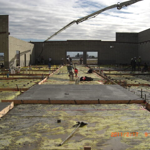 A low-angle photograph shows a large concrete floor pour in progress at a construction site. A long, articulated concrete pump hose extends from the top right of the frame, delivering a fresh mix of concrete onto a prepared surface. The floor has been framed with wooden forms and rebar, with a yellow vapor barrier visible. Workers in hard hats and vests are scattered across the site, using tools to level and smooth the concrete. The partially completed walls of the building, made of concrete masonry units (CMU), are visible in the background. A timestamp in the bottom right corner reads "2011/2/27 9:32am".
