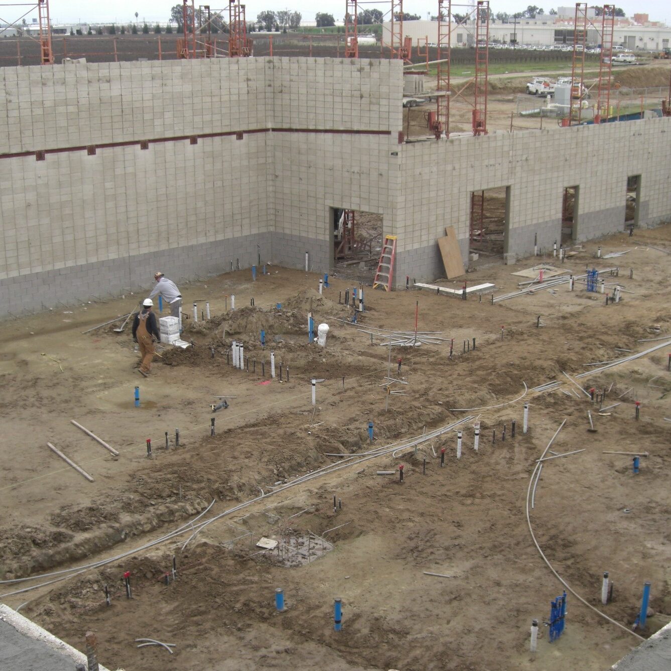 A wide-angle, slightly elevated shot of a large building under construction. The foundation is a patch of muddy, light-brown dirt with several workers in hard hats and safety vests installing extensive plumbing and electrical conduit. The walls are made of gray concrete blocks with several rectangular openings for windows or doors. Scaffolding is visible along the top of the walls. The scene is shot under a gray, overcast sky.