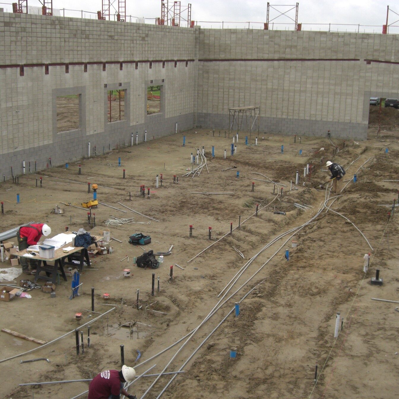 A wide-angle, slightly elevated shot of a large building under construction. The foundation is a patch of muddy, light-brown dirt with several workers in hard hats and vests installing extensive plumbing and electrical conduit. A long, gray wall made of concrete blocks with a few rectangular openings for windows or doors curves around the perimeter. The top of the wall is outlined in a thin red line, and rebar sticks out of the top. Construction equipment is visible in the background and on the left.