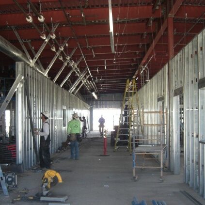 A low-angle, eye-level shot from inside a long hallway under construction. The walls are framed with exposed metal studs, and a red steel grid is visible on the ceiling. Several construction workers are visible, along with tools and equipment, including a yellow ladder and a saw. The hallway leads to a brightly lit opening at the other end.