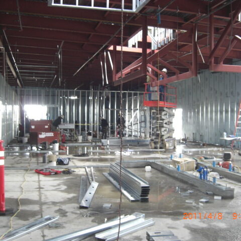 A wide-angle photograph shows the interior of a building under construction. The structure's steel frame is visible, with red beams forming the ceiling and roof supports. The walls are framed with galvanized steel studs. The concrete floor is wet and covered in debris, including tools, equipment, and metal framing materials. In the background, workers are visible on ladders and a scissor lift, engaged in various construction tasks. The lighting is bright, possibly from natural light entering through an open section of the building. A digital timestamp in the bottom right corner of the image reads "2011/4/8 9:57am".