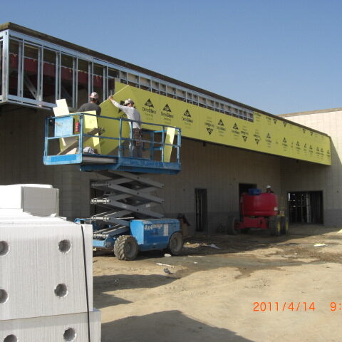 A daytime construction photograph shows two workers in a blue scissor lift, applying yellow sheathing to the exterior of a building. The sheathing is branded with the logos of "DensGlass" and "Georgia-Pacific". The building's walls are made of concrete masonry units (CMU), and a steel-framed canopy or overhang extends from the structure. A red lift is visible in the background, and construction materials are scattered on the dirt ground in the foreground, including a pallet of what appears to be concrete blocks. The sky is bright and clear. A timestamp at the bottom right corner reads "2011/4/14 9:10am".