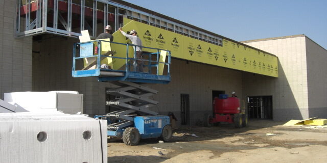 A daytime construction photograph shows two workers in a blue scissor lift, applying yellow sheathing to the exterior of a building. The sheathing is branded with the logos of "DensGlass" and "Georgia-Pacific". The building's walls are made of concrete masonry units (CMU), and a steel-framed canopy or overhang extends from the structure. A red lift is visible in the background, and construction materials are scattered on the dirt ground in the foreground, including a pallet of what appears to be concrete blocks. The sky is bright and clear. A timestamp at the bottom right corner reads "2011/4/14 9:10am".