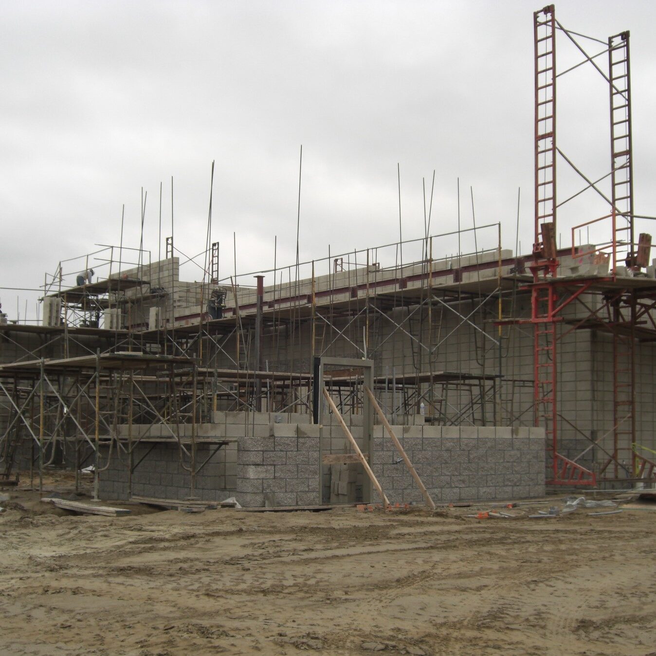 A wide-angle, eye-level shot of a two-story building under construction on a cloudy day. The building's walls are made of gray cinder blocks and are surrounded by extensive scaffolding and ladders made of red and gray metal. Piles of dirt and construction materials are scattered across the muddy ground in the foreground. In the left background, large red and blue pipes are visible.