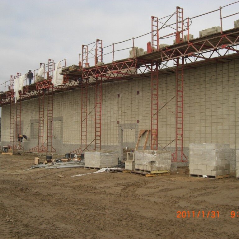 A construction photograph shows the exterior of a large, single-story building under construction. The wall is made of light-colored concrete masonry units (CMU). Red-colored metal scaffolding is set up along the entire length of the wall, with workers visible on the elevated platform. Pallets of CMU blocks, wrapped in plastic, are stacked on the dirt ground in front of the building. The ground is a mix of dirt and mud with tire tracks. The sky is overcast and grey. A timestamp in the bottom right corner reads "2011/1/31 9:52am".