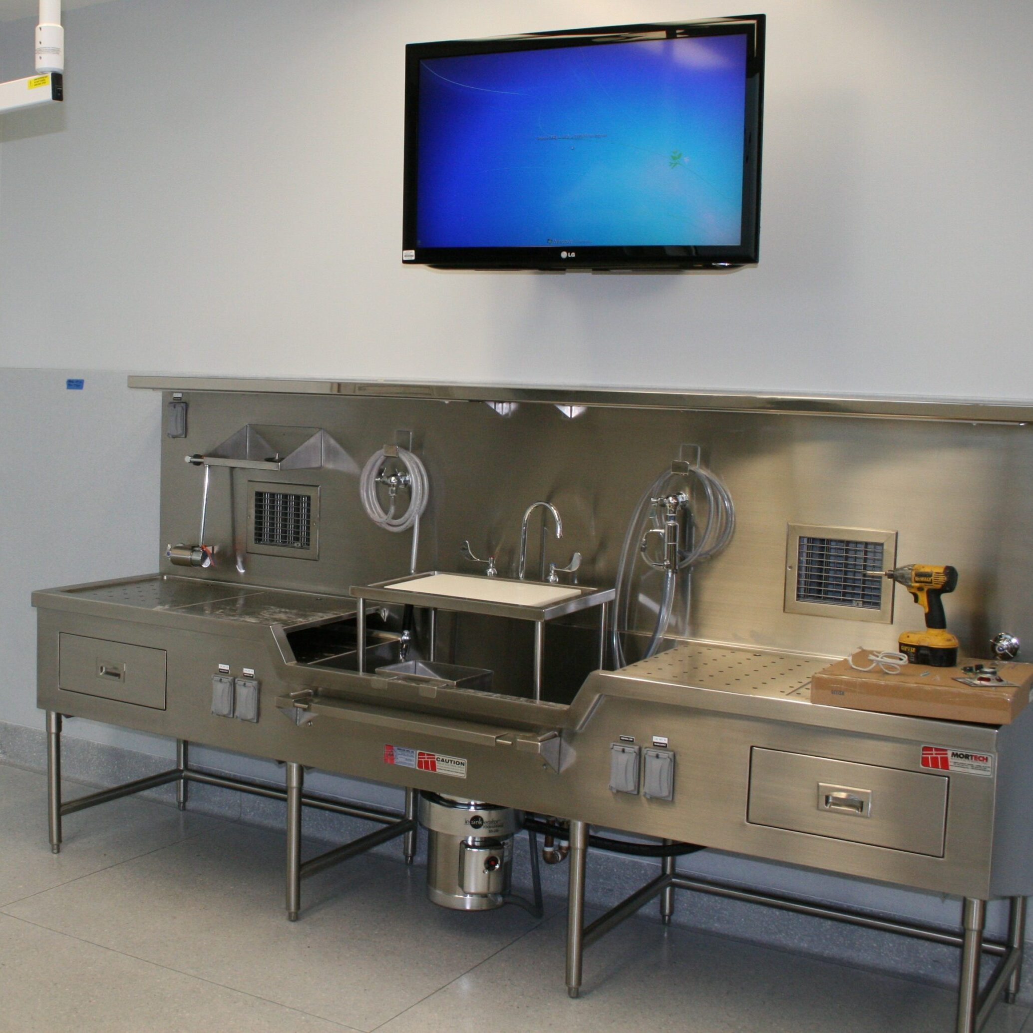 A low-angle, eye-level shot of a large, stainless steel autopsy and dissection sink unit. The unit has two large sinks, one of which is equipped with a food disposal. It features a variety of faucets, hoses, and an overhead lighting fixture. A power drill and goggles sit on the counter of the right sink. A large, dark-screen monitor is mounted on the wall above the unit, with a visible Windows desktop. The room has light gray walls and a pale-gray tiled floor.