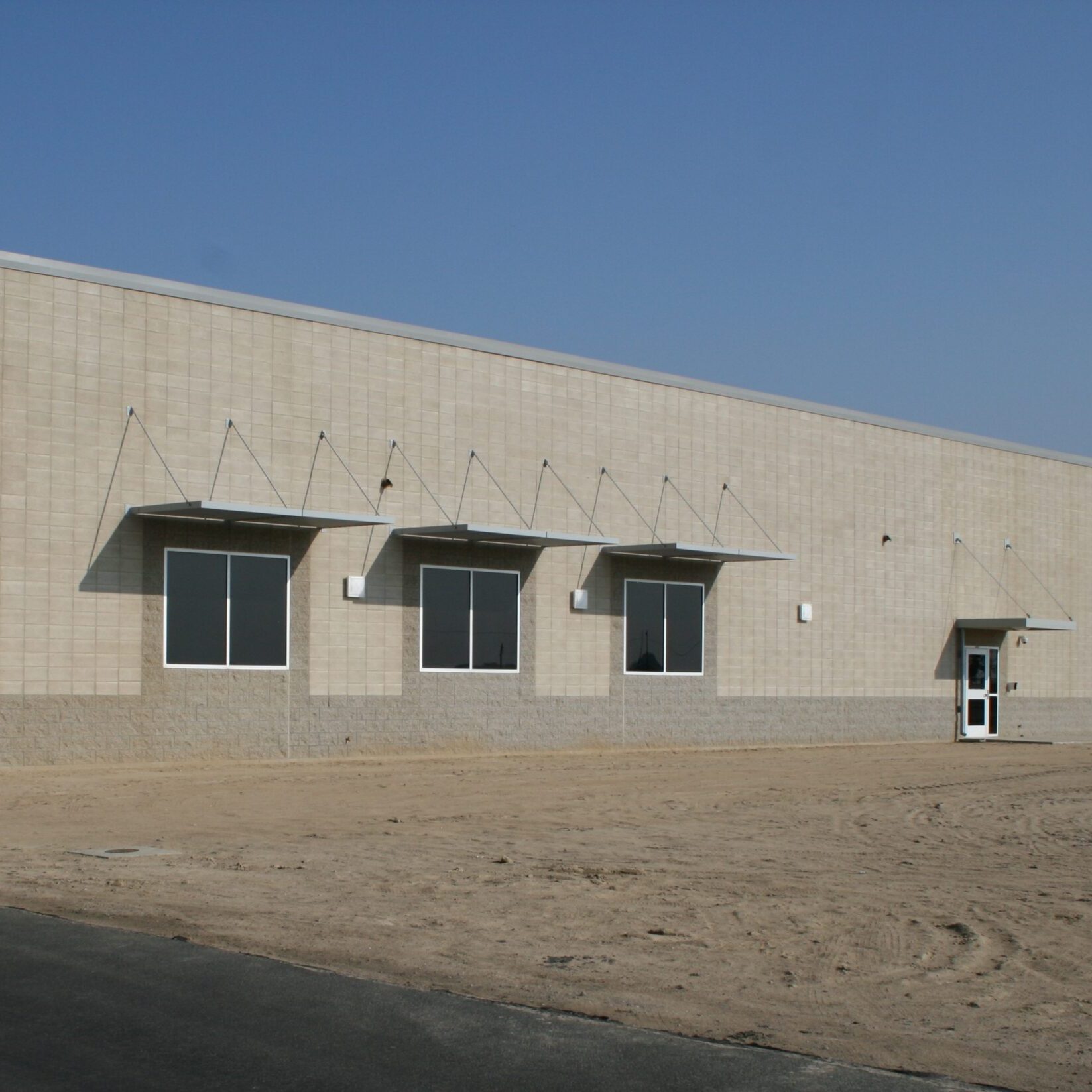 A low-angle, eye-level shot of the side of a large, modern, institutional-style building. The walls are made of two different-colored concrete blocks: a lighter beige on top and a darker gray on the bottom. Several rectangular windows with gray metal canopies are visible. A single door is on the right side of the building. The foreground is a dirt and gravel lot, with a paved road marked with "No Parking" on the bottom left. The sky is bright blue and cloudless.