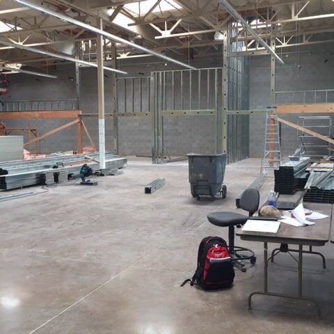 An interior construction site showing the concrete floor and exposed steel framing for new walls. Rolls of insulation and stacks of metal studs are on the floor. A ladder, trash can, and work tables are also visible.